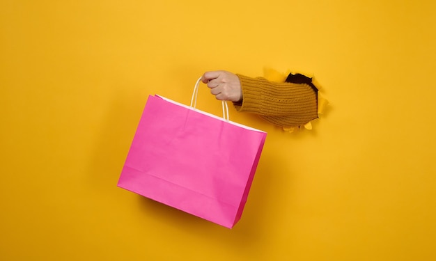 Female hand with a pink paper shopping bag sticking out of a torn hole in a yellow paper background. Refusal from plastic, start of sales