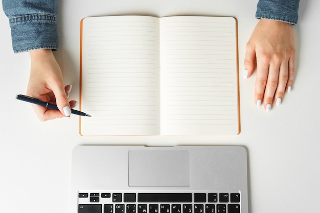 Female hand writing in a notebook on table with laptop, top view