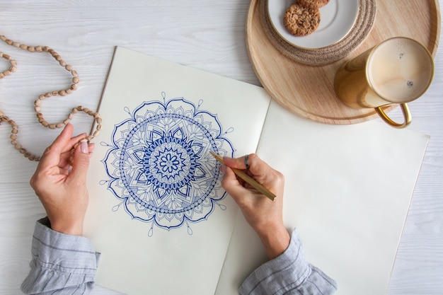 Female hands close up draw Decorative round floral mandala. Hobby and home relaxation. A mug of coffee and cookies on a wooden tray. White background.