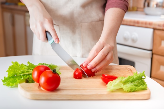 Female hands cut a juicy red tomato into slices with a knife on a wooden cutting board. A method of preparing vegetables and ingredients before cooking.