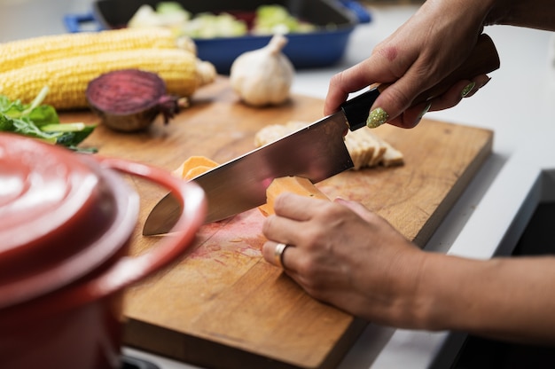 Female hands cutting sweet potato on a wooden kitchen board full of various autumn vegetables.