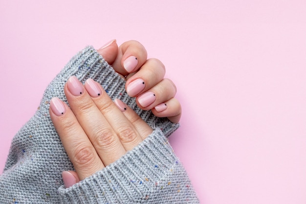 Female hands in a gray knitted sweater with trendy beautiful manicure - pink nude nails with black small dots on a pink background with copy space