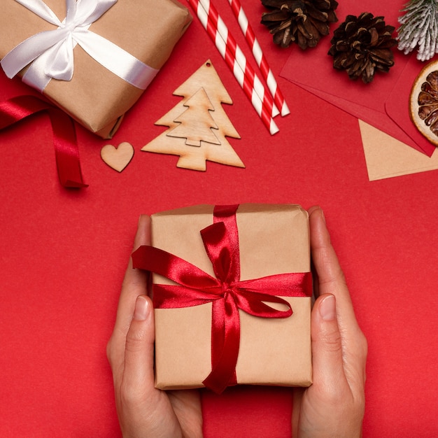 Female hands hold christmas gift box on red background with decoration top view. Holiday background,