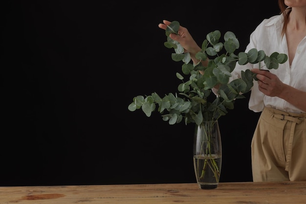 Female hands hold eucalyptus in glass vase on wooden table dark background mockup