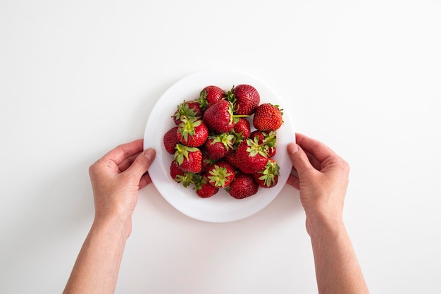 Female hands hold a plate with strawberries on a white table background Top view flat lay