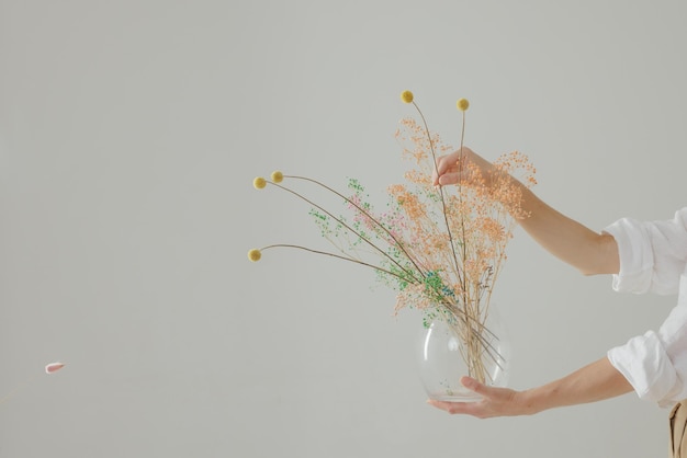 Female hands hold a transparent glass vase with dried flowers on a light background