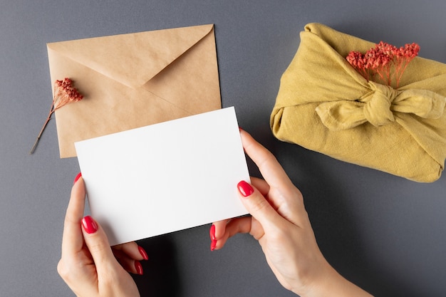 Female hands holding blank paper card over gray table with craft envelope furoshiki gift