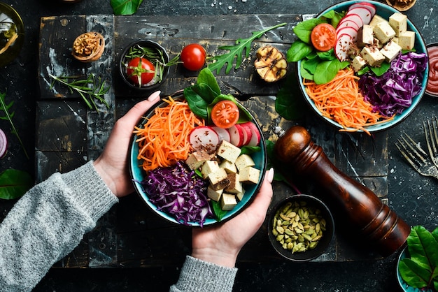 Female hands holding a bowl of vegan lunch tofu cheese spinach cabbage radishes and carrots The concept of healthy eating Top view