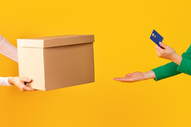 Female hands holding credit card and taking cardboard box from carrier or seller on yellow studio