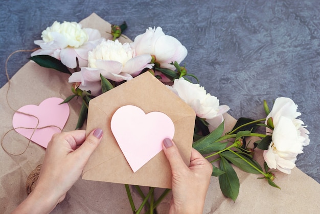 Female hands holding envelope with pink paper heart, sending flowers and invitation for romantic event