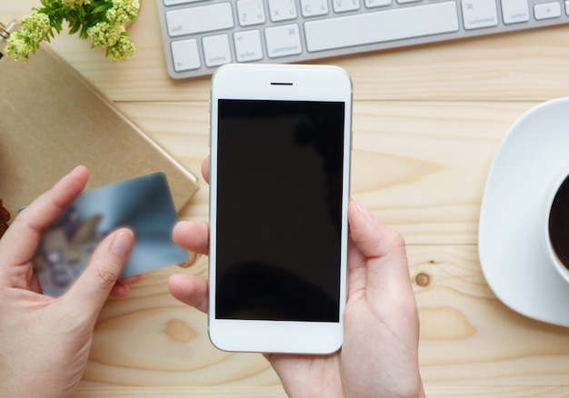 Female hands holding phone with blank screen and a credit card over the desk 