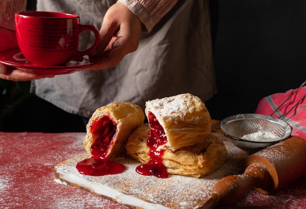 Female Hands Holding Red Cup of coffee near Puff staffed with plum or red currant jam on the table