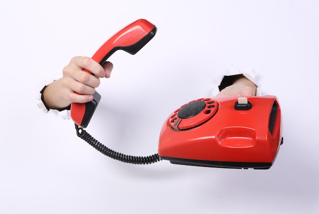 Female hands holding retro rotary phone through torn hole white paper