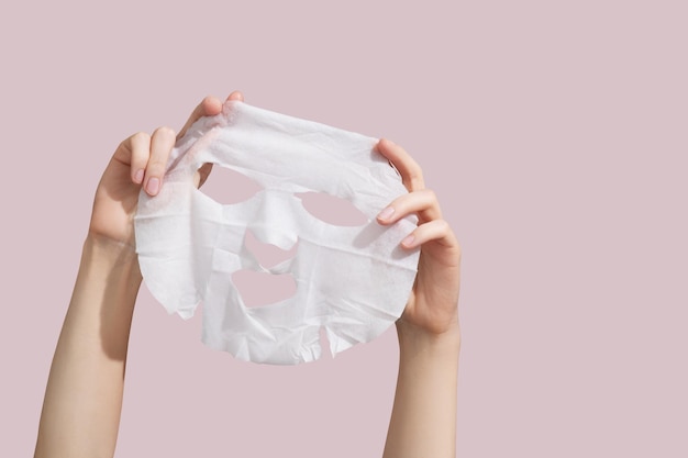 Female hands holding sheet of white mask on pink background