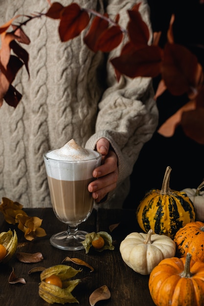 Female hands holding steaming cappuccino, latte coffee with pumpkins, autumn leaves on wooden table