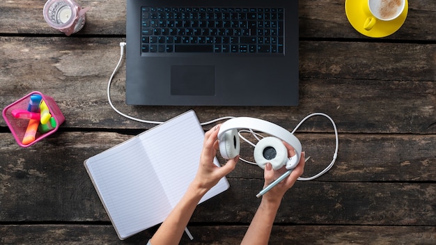 Female hands holding white headphones plugged into laptop computer next to an open notebook