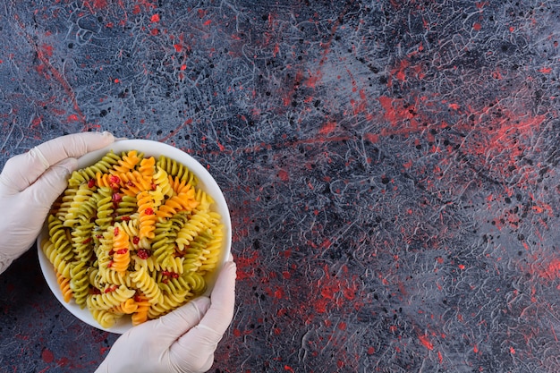 Female hands holding a white plate of raw dry multi colored Fusilli pasta on a dark surface 