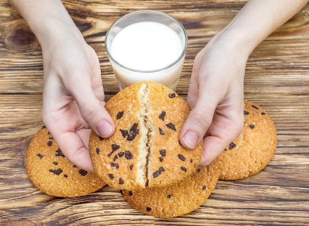 Female hands holds chocolate chip cookie on background of wooden table with glass of milk and cookies