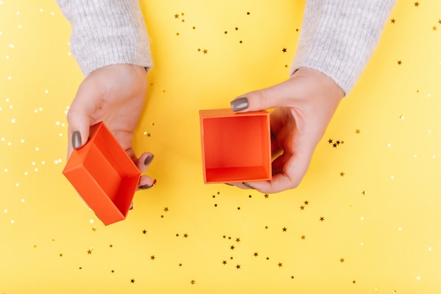 Female hands open a red gift box. Yellow background with gold sequins. Concept of the holidays