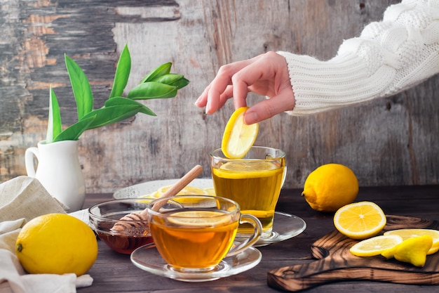 Female hands put a slice of lemon into a cup of tea, lemons and a bowl of honey on a wooden table. Vitamin Organic Nutrition