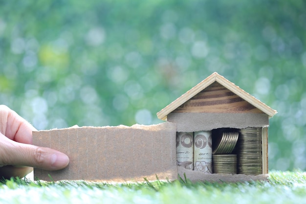 Female hands rip a brown sheet of paper and money in model house on natural green backgroundBusiness investment and real estate concept