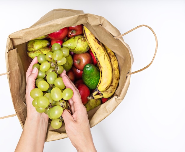 Female hands take out a bunch of grapes from a paper bag full of different fruits isolated on a white background Delivery and purchase of healthy eco food