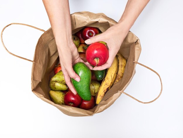 Female hands take out a red apple and avocado from a paper bag full of different fruits isolated on a white background Delivery and purchase of healthy eco food