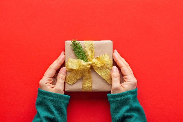 Female hands in turquoise sweater holding Christmas present in paper wrap on red background, top view