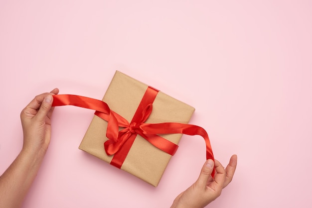 Female hands untie a red ribbon on a gift wrapped in brown kraft paper