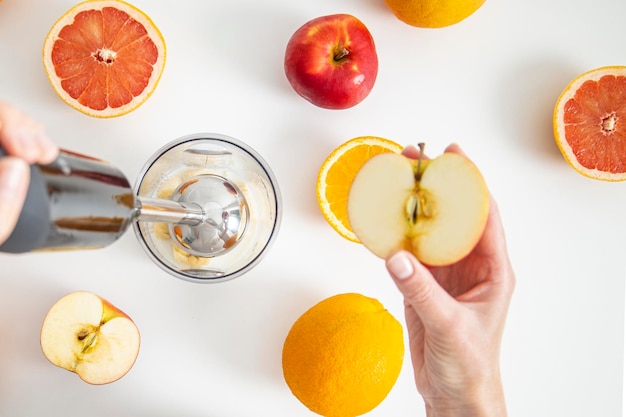 Female hands use a hand blender to mix fresh fruits to make a diet smoothie on a white background Top view flat lay