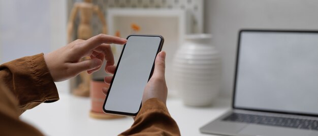 Female hands using mock up smartphone while sitting at worktable with mock up laptop in home office