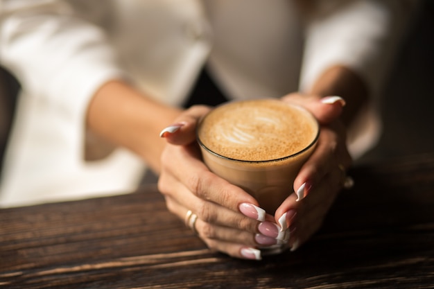 Female hands with beautiful manicure close-up hold a cup with hot coffee on a wooden table.