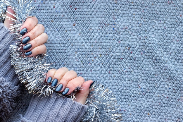 Female hands with dark gray blue glittered nails on knitted background with silver Christmas tinsel