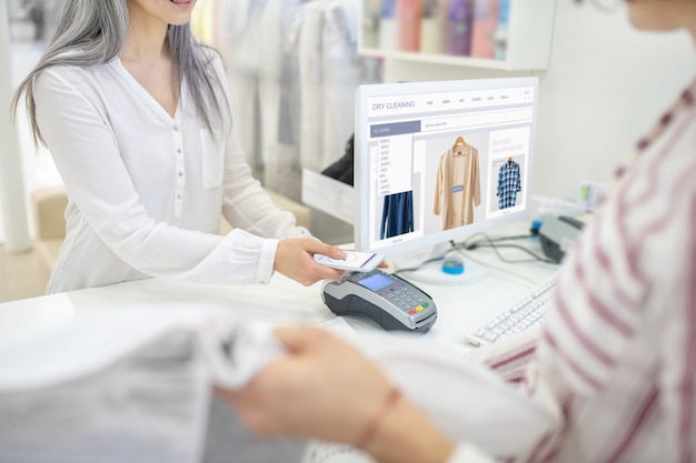 Female hands with smartphone over pos terminal on counter near computer screen and seller