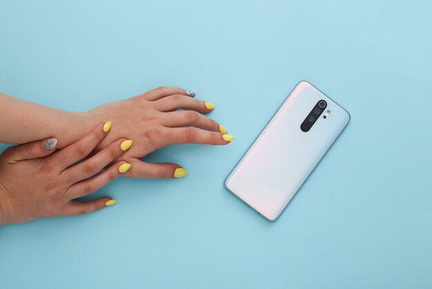 Female hands with a yellow manicure and smartphone on a blue