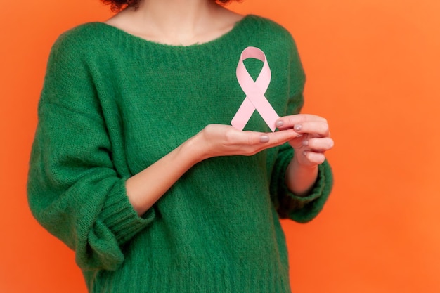 Female health check awareness. Woman hair in green casual style sweater holding pink ribbon, symbol of breast cancer, support of oncology patients. Indoor studio shot isolated on orange background.