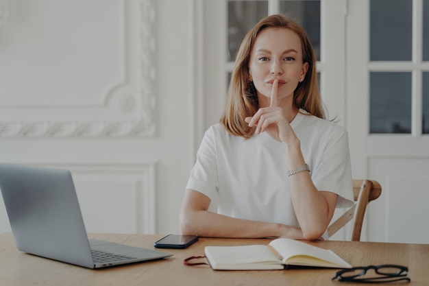 Female hold finger on lips showing silence gesture asking to be quiet sitting at desk with laptop