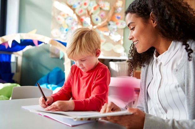 Female infant school teacher working one on one with a young white schoolboy sitting at a table in a classroom writing close up