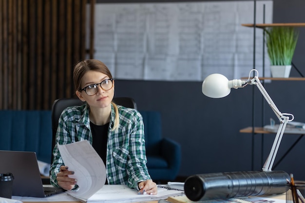 Female interior designer working in office with laptop. Architect thinks over architectural plan, searching new ideas for construction project. Woman sitting at workplace. Business portrait concept.