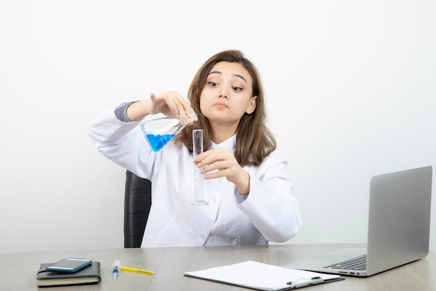 Female laboratory researcher sitting at the desk and holding medical glass bottle with blue liquid . High quality photo