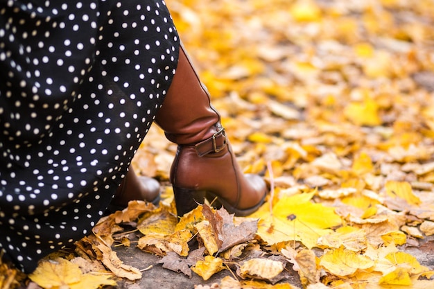 Female legs in long brown leather boots and a long skirt against the background of yellow leaves Horizontal photo