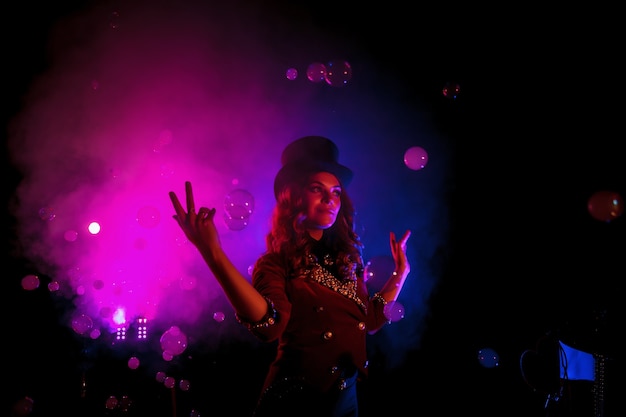 Female magician, an illusionist in theatrical clothes, makes show with soap bubbles on black background. Man actress in stage costume and top hat on her head. Concept of theatrical performance