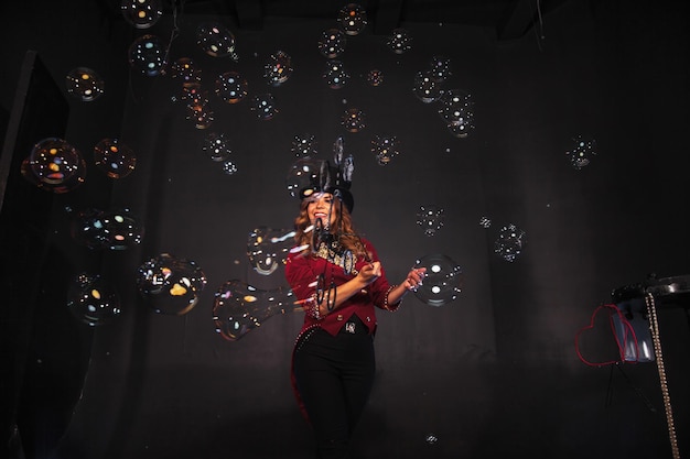 Female magician makes with soap bubbles show an illusionist in theatrical clothes on black background Woman actress in stage costume Concept of theatrical performance and fun show Copy space