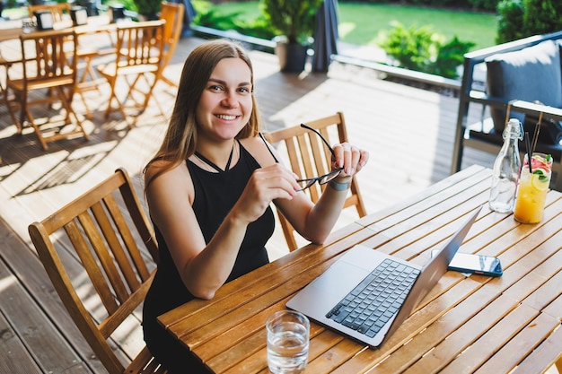 Female manager freelancer enjoying coffee break on summer terrace of cozy cafe working remotely on laptop smiling looking at laptop Work online in the summer