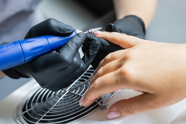 Female master using electric nail file while doing manicure for client in beauty salon