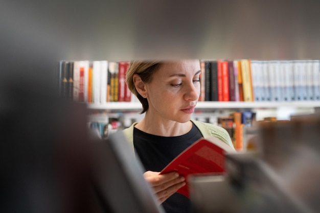 Female mature student looking for book at library bookshelf studying at university campus or college