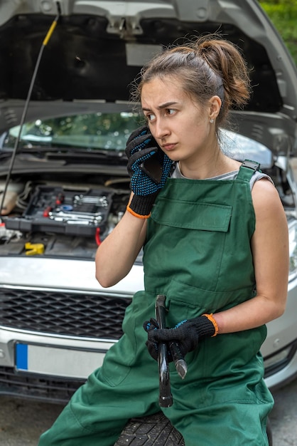 A female mechanic in overalls is resting near a broken car and waiting for help from a partner