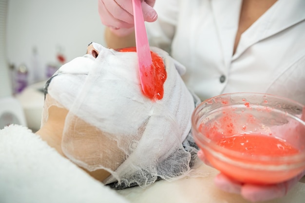 Female medical worker applying on face red alginate mask her client in beauty salon