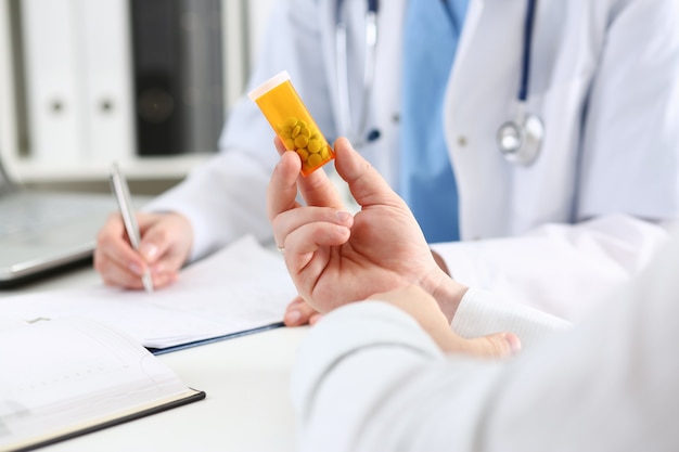 Female medicine doctor writes prescription at worktable while patient hand hold jar of pills.