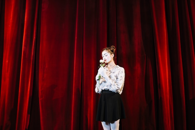 Female mime standing in front of red curtain smelling white rose